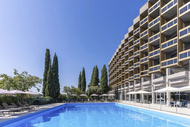 Daytime outdoor pool with cypress trees, umbrellas and the hotel building
