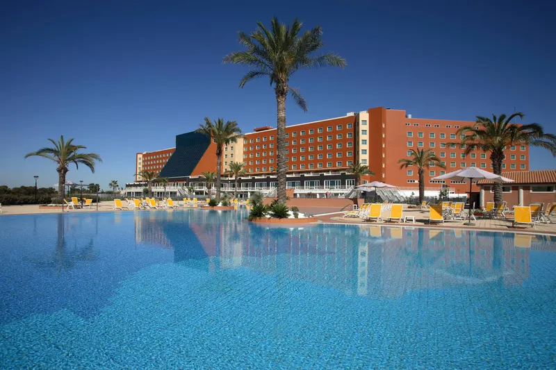 Hotel exterior seen across the large blue pool with palm trees